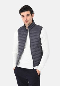 Gilet imbottito senza maniche di colore grigio scuro, con colletto alto e motivo a cuciture orizzontali, abbinato a una camicia bianca a maniche lunghe.