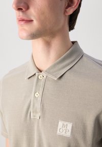 Beige polo skjorte laget av strukturert stoff, med to knapper foran, brodert logo på brystet, og klassisk kragedesign.