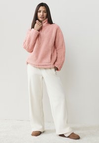 Maglione rosa e peloso con colletto alto e mezza cerniera, abbinato a pantaloni crema a gamba larga e scarpe marroni senza lacci. Materiali morbidi e texturizzati.