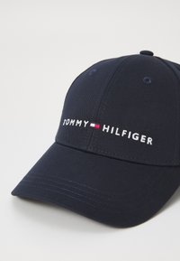 Kleitas jūras zila beisbola cepure, kas izgatavota no auduma. Satur baltu izšuvumu tekstu "TOMMY" un "HILFIGER" ar sarkanu un baltu akcentu līniju. Ar izliekto vizu.