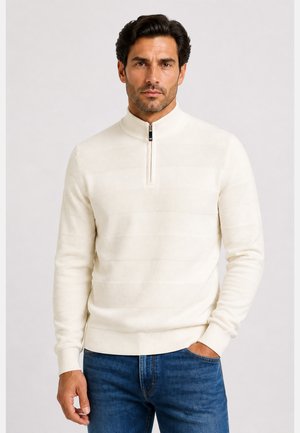 Mann mit dunklen Haaren, der einen weißen texturierten Halb-Zip-Pullover und blaue Jeans trägt, steht mit einer Hand in der Tasche vor einem einfarbigen Hintergrund.