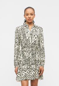 Γυναίκα με πλεξούδες που φοράει φόρεμα μακρυμάνικο σε ασπρόμαυρο μοτίβο με ζώνη στη μέση, στέκεται μπροστά σε απλό φωτεινό φόντο.