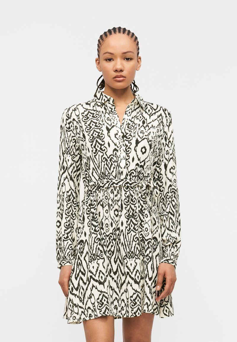 Γυναίκα με πλεξούδες που φοράει φόρεμα μακρυμάνικο σε ασπρόμαυρο μοτίβο με ζώνη στη μέση, στέκεται μπροστά σε απλό φωτεινό φόντο.