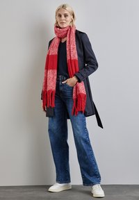 Cappotto trench navy, sciarpa a scacchi rossi, top scuro, jeans blu a gamba larga e sneakers bianche. La sciarpa presenta delle frange, mentre i jeans hanno una texture sbiadita.