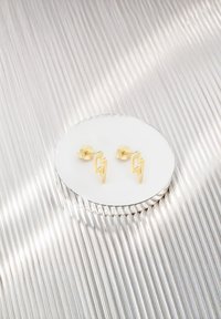 Orecchini a stud a forma di fulmine dorato esposti su uno specchio circolare, con texture lisce e un design elegante e moderno.