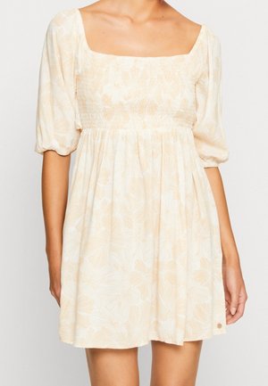 Femme portant une robe courte fleurie beige avec manches bouffantes et encolure carrée, vue des épaules à mi-cuisse.