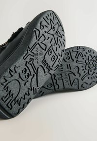 Schwarze Gummisohle mit eingravierten verspielten Designs, darunter Sterne, Herzen und verschiedene Figuren, mit einer strukturierten Oberfläche für besseren Halt.