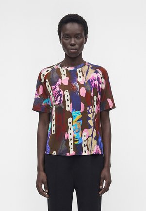 Femme portant une chemise à manches courtes avec un imprimé abstrait coloré aux tons marron, rose, bleu et crème, associée à un pantalon noir, debout sur un fond blanc.