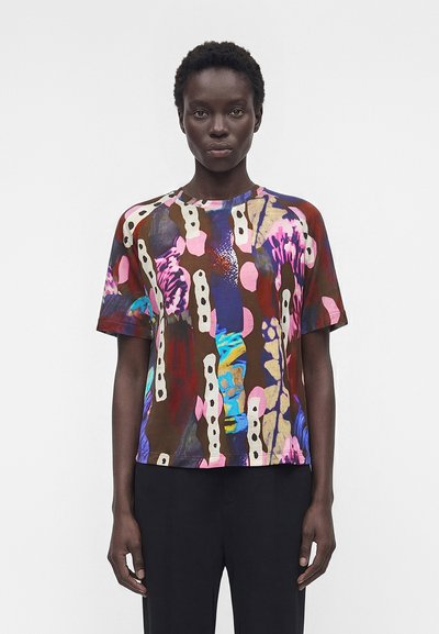 Mujer con una camiseta de manga corta con estampado abstracto y colorido en tonos marrón, rosa, azul y crema, combinada con pantalones negros, de pie sobre un fondo blanco.