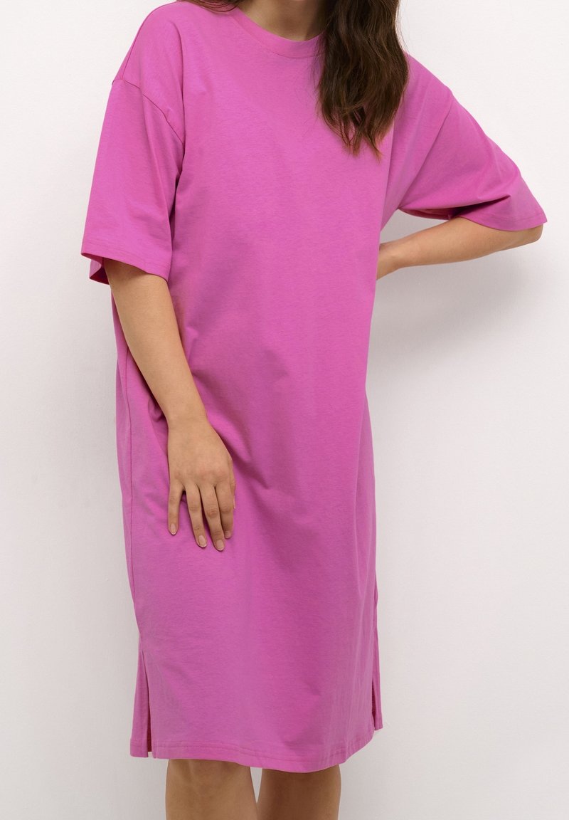 Femme portant une robe rose ample à manches courtes et longueur genou, debout, une main sur la hanche et l'autre reposant le long du corps.