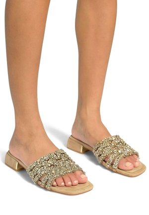 Sandali slide beige con accenti di cristallo, dotati di tacco piatto e design open-toe. Parte superiore testurizzata con decorazioni decorative.
