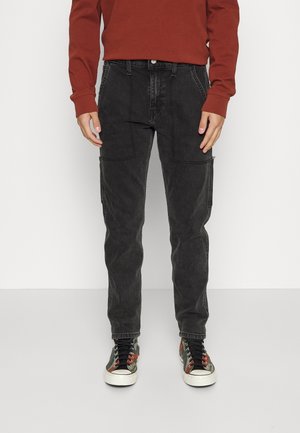 Personne portant un jean cargo noir, des baskets camouflage et un t-shirt orange brûlé à manches longues, debout devant un fond uni.