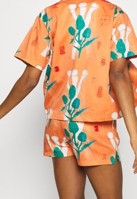 Pomarańczowa bawełniana bluzka z krótkim rękawem i dopasowane szorty z zielonymi wzorami roślinnymi i abstrakcyjnymi motywami. Luźny krój, lekka tekstura.