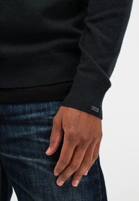 Close-up van een donkergekleurde hand met een zichtbare litteken op de rug, gekleed in een zwarte trui en blauwe spijkerbroek.