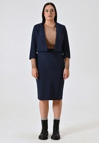 Navy blauwe cropped blazer gecombineerd met een nauwsluitende kokerrok. Het model draagt een beige top en zwarte veterschoen enkellaarsjes. Gladde stof, op maat gemaakte ontwerp.