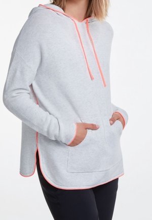 Kvinde iført lysegrå hættetrøje med lyserød kant og snore, hænderne i forlomme, kombineret med sorte bukser mod hvid baggrund.