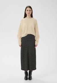 Pull beige en tricot à coupe décontractée associé à une jupe midi noire à pois avec de grands pois dorés et des bottes noires montant jusqu'au genou.