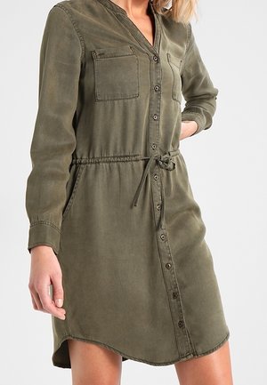 Femme portant une robe boutonnée vert olive, manches longues, poches poitrine, taille à cordon et poches latérales, debout sur fond blanc.