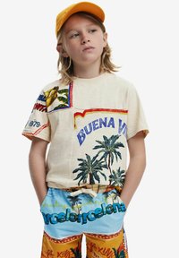 Beige T-shirt med färgglada broderade lappar i form av palmträd och text; matchad med blå och orange mönstrade shorts.