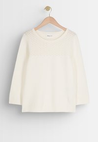 Bílý pletený svetr s texturovaným vzorem na horní části, zaobleným výstřihem a tříčtvrtečními rukávy, zavěšený na dřevěném ramínku.