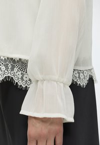 Manche de blouse blanche transparente avec poignet élastique et détail de bordure en dentelle sur tissu noir, main détendue sur le côté.