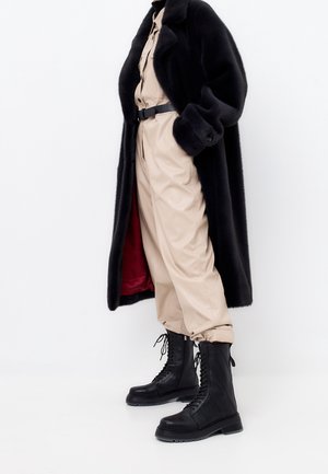 Person, die einen beigen Overall mit Gürtel, einen langen schwarzen Pelzmantel mit rotem Futter und schwarze Schnür-Kampfstiefel vor weißem Hintergrund trägt.