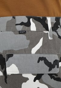 Gros plan sur un pantalon camouflage gris, noir et blanc avec une poche cargo et une petite étiquette noire de la marque "HUF" près de la taille.