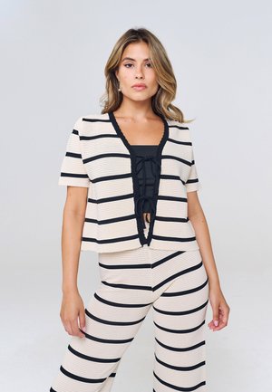 Femme portant un haut à manches courtes rayé horizontalement crème et noir et un pantalon large assorti, debout avec une expression neutre sur un fond uni.