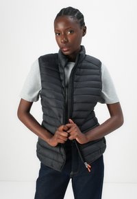 Black puffer vest with a high collar, quilted design, and zip closure. Worn over a gray t-shirt, features orange zipper details.