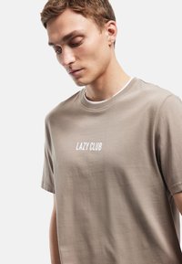 Lysebrun bomulds t-shirt med rund halsudskæring og korte ærmer. "LAZY CLUB" trykt i hvidt tværs over brystet. Glat tekstur.