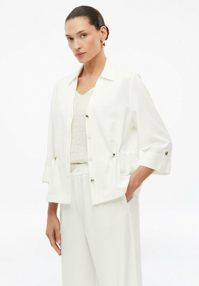 Woman modeling loose white buttoned jacket, knit top, and wide-leg pants, with hair slicked back and gold hoop earrings, on white background.