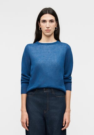 Kvinde med langt mørkt hår iført gyldne øreringe, en blå sweater og mørke højtaljede jeans, stående mod en ensartet lys baggrund.
