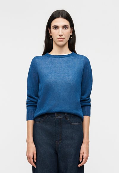 Femme aux longs cheveux foncés portant des boucles d'oreilles anneaux dorés, un pull bleu et un jean taille haute foncé, debout devant un fond clair uni.
