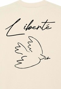 Beige t-krekls ar melnu līniju zīmējumu, kurā attēlota baloža un vārds "Liberté" cursīvu fontā virs tā. Vienkāršs, minimālistisks dizains.