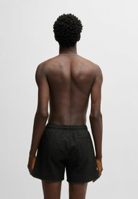Costumi da bagno neri con texture, dotati di una cintura elastica e tasca laterale, mostrati su una persona vista da dietro.
