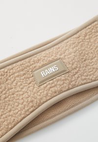 Close-up van beige fleece stof met gestikte randen en een rechthoekig beige label met de opdruk "RAINS" in witte letters.