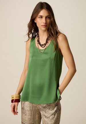 Donna con una camicetta verde senza maniche, pantaloni beige con motivo, collane a strati e braccialetti spessi, in piedi con una mano in tasca.