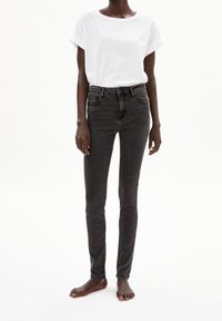 Personne portant un t-shirt blanc uni à manches courtes et un jean skinny noir taille haute, debout pieds nus sur un fond blanc.