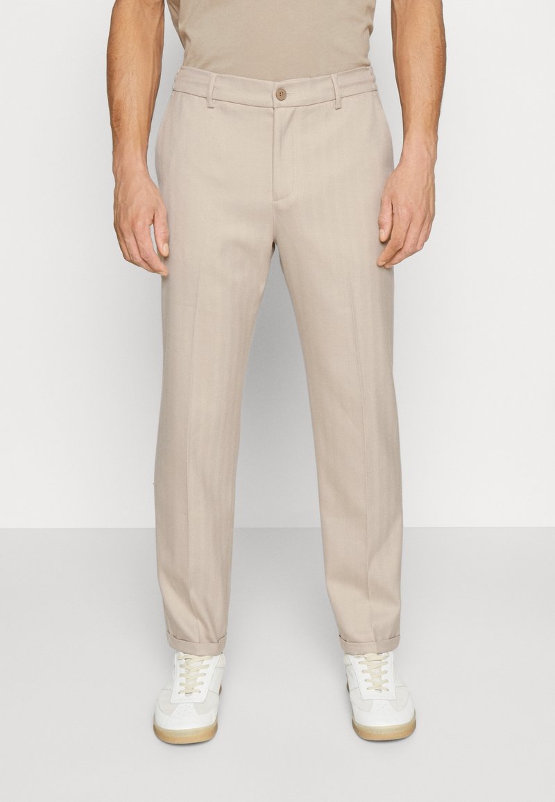 Homme portant un pantalon beige droit avec des ourlets retroussés et des baskets blanches, debout devant un fond clair uni.