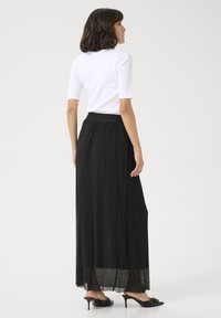 Jupe maxi plissée noire avec une taille ajustée, associée à un haut blanc à manches courtes. Le mannequin porte des mules à talons noirs. Design simple.