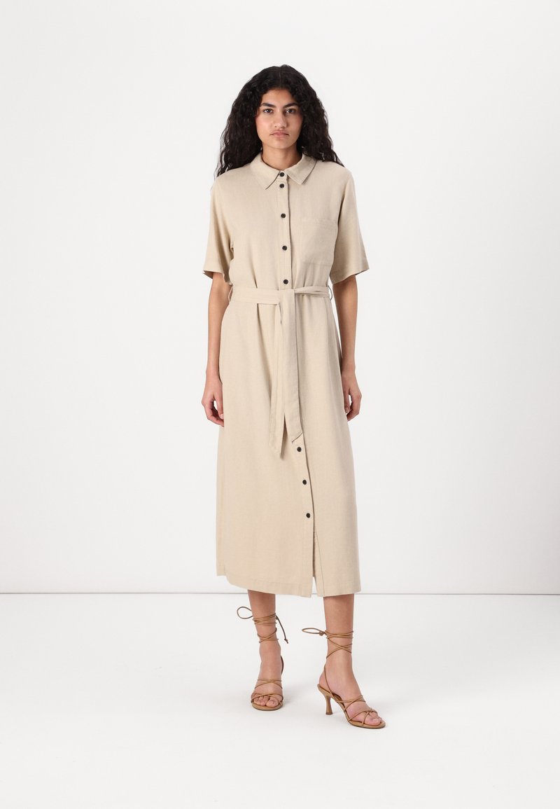 Abito button-up in misto lino beige con maniche corte, cintura con nastro e tasca frontale, abbinato a sandali con tacco alto beige con lacci.
