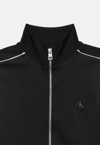 Čierna zipová bunda s vysokým golierom, rebrovanou textúrou, strieborným zipsom a diskrétnym logom na prednej strane. Hladká tkanina s pásikovaním.
