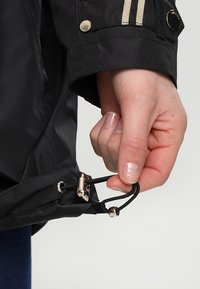 Giacca impermeabile nera con coulisse in vita, cerniera con dettagli dorati e inserti a righe sulle maniche.