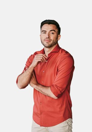 Mann mit kurzen dunklen Haaren und Bart, trägt ein hochgekrempeltes, rotes Hemd und beige Hosen, steht mit einer Hand in der Nähe seines Kragens.