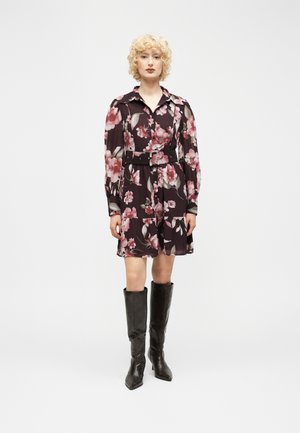 Kobieta o kręconych blond włosach, ubrana w czarną sukienkę w kwiaty z paskiem i czarne kozaki sięgające kolan, stoi na tle jednolitego jasnego tła.