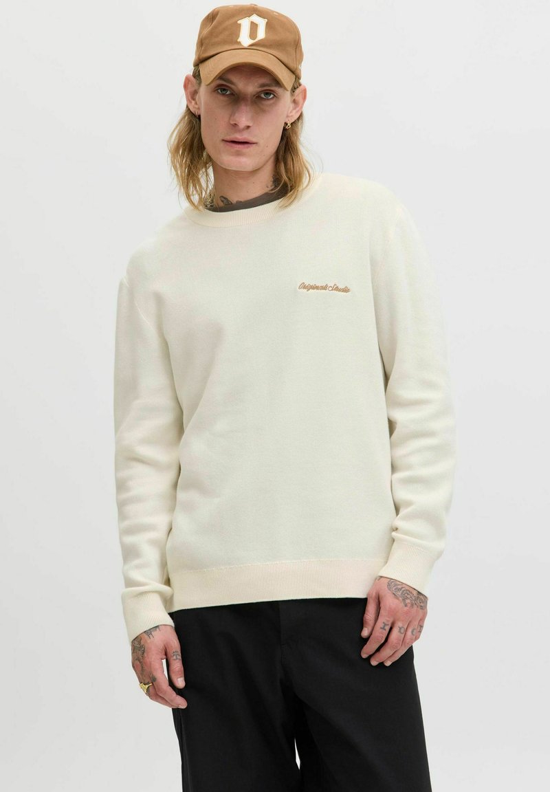 Cremefarbener Sweatshirt mit gerippten Bündchen und Saum, mit einem hellbraunen gestickten Logo auf der Brust. Kombiniert mit dunkler Hose.