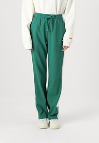 Zaļas ribiņmēteļa bikses ar stringu viduklī, baltas sānu malas un logotipa uzšuvums. Saskaņotas ar baltām sporta kurpēm un vieglu džemperi.