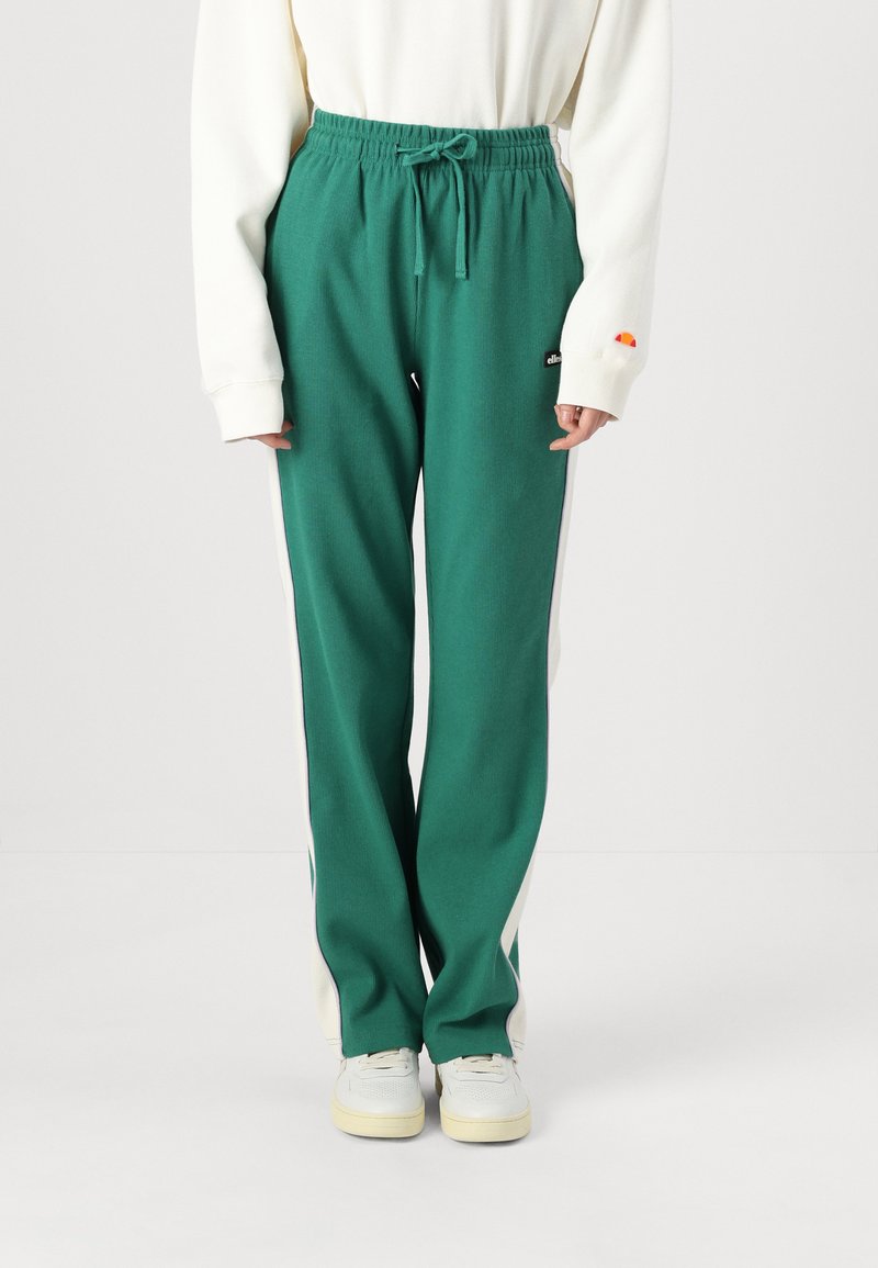 Zaļas ribiņmēteļa bikses ar stringu viduklī, baltas sānu malas un logotipa uzšuvums. Saskaņotas ar baltām sporta kurpēm un vieglu džemperi.