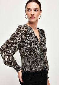 Blusa nera con pois bianchi, dotata di scollo a V, vita arricciata e maniche lunghe a sbuffo. Tessuto leggero e trasparente che aggiunge texture.