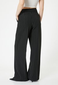 Pantalons larges noirs avec des rayures blanches, taille haute et poches latérales. Le tissu a une texture lisse. Les ourlets sont légèrement évasés.